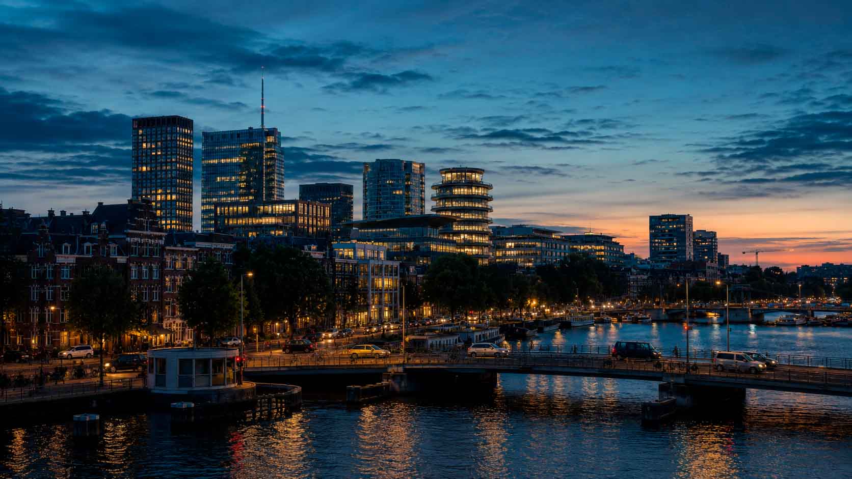 Amsterdam waterfront at dusk with illuminated office buildings, bridges, and canal reflections, used as a regional context image for data center photographer services.